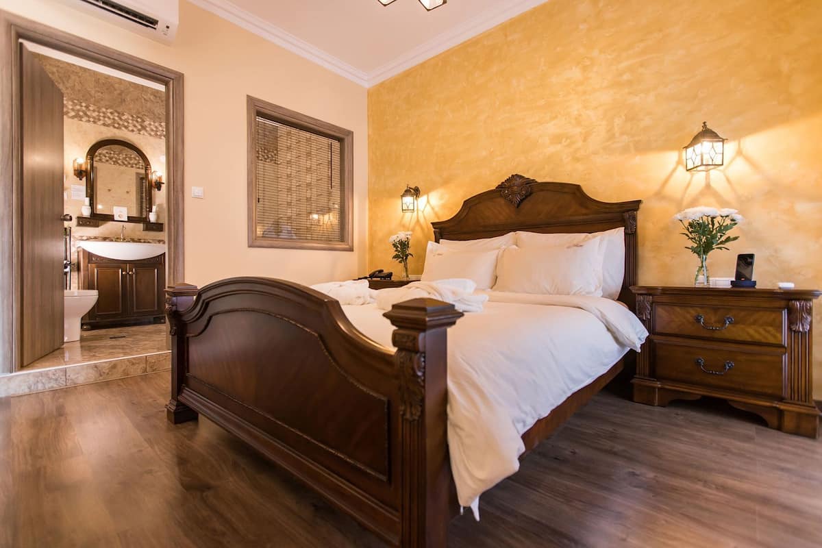 A bedroom with a wooden bed, a nightstand, a bedside table, a mirror, a window with blinds, and a bathroom visible through an open door.