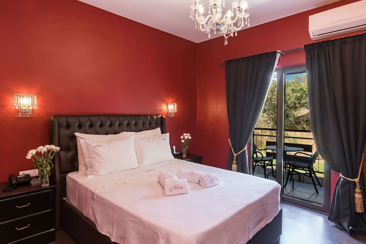 A hotel room with a large bed, a chandelier, a balcony with outdoor seating, and an air conditioning unit.