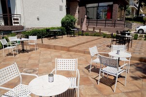 Un patio al aire libre con sillas y mesas de metal blanco, una pequeña fuente y un edificio con fachada de piedra al fondo.