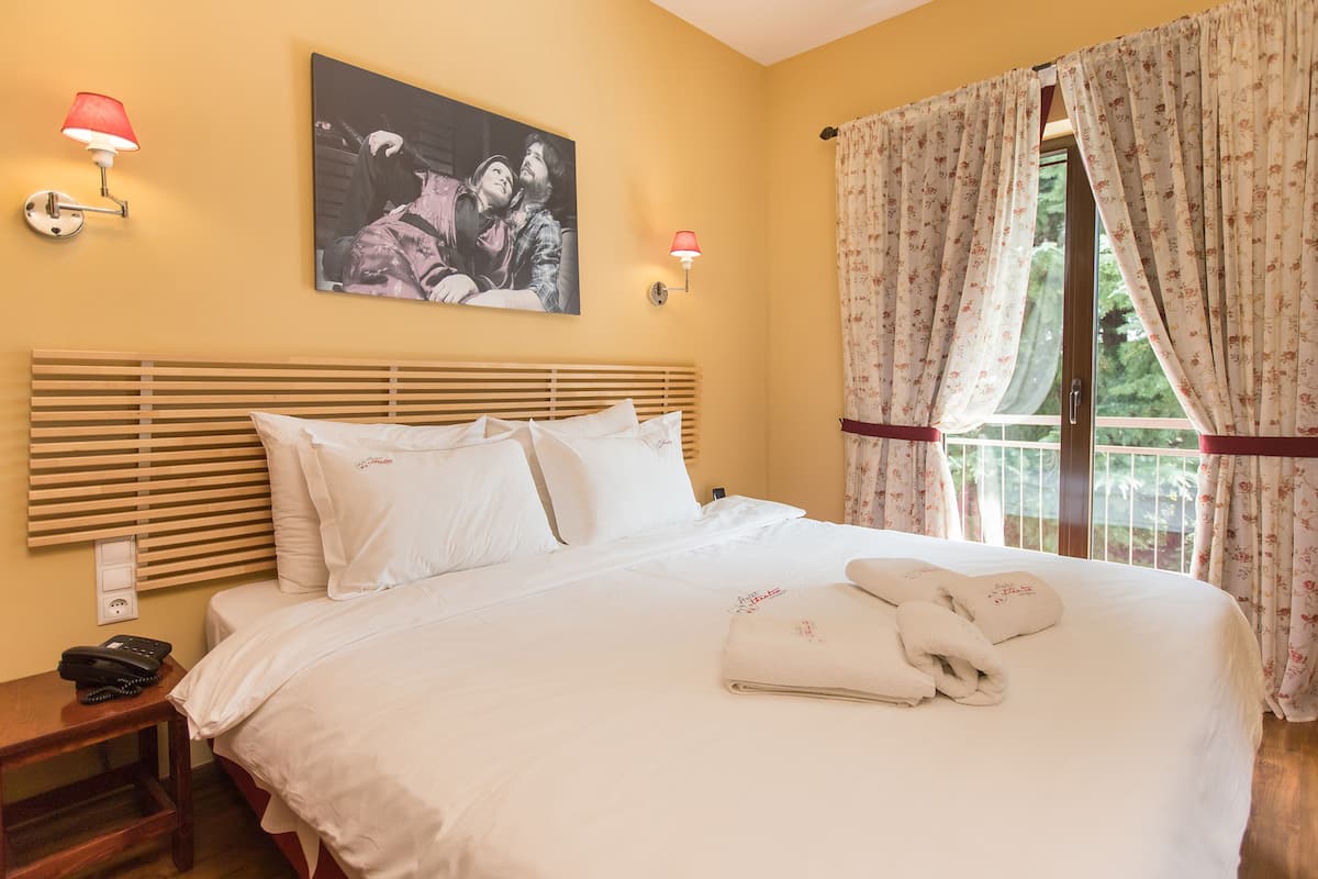 A neatly made bed with white linens, a wooden headboard, and a painting above it. There are two wall-mounted lamps with red shades, and a window with floral curtains.