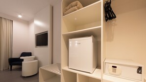 Una habitación de hotel moderna con refrigerador, televisor de pantalla plana, zona de estar con una silla y una mesita, y perchero.