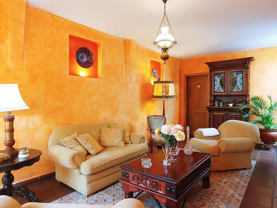 Una sala de estar con un sofá, sillones, una mesita de centro y una lámpara. Se observa una puerta con un vitral, una planta en maceta y un extintor en la pared.