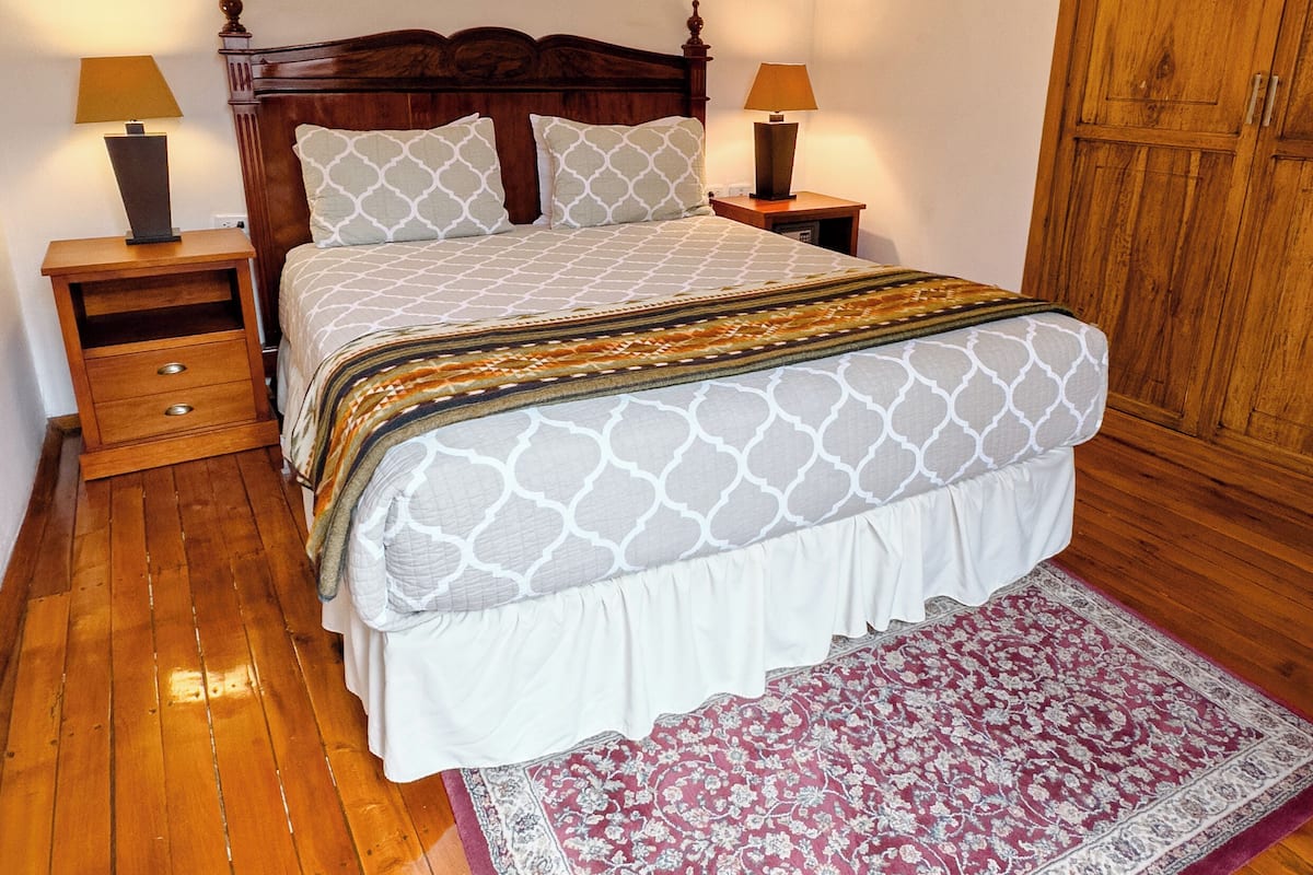 A bedroom with a wooden bed, two bedside tables with lamps, a wooden wardrobe, and a patterned rug.