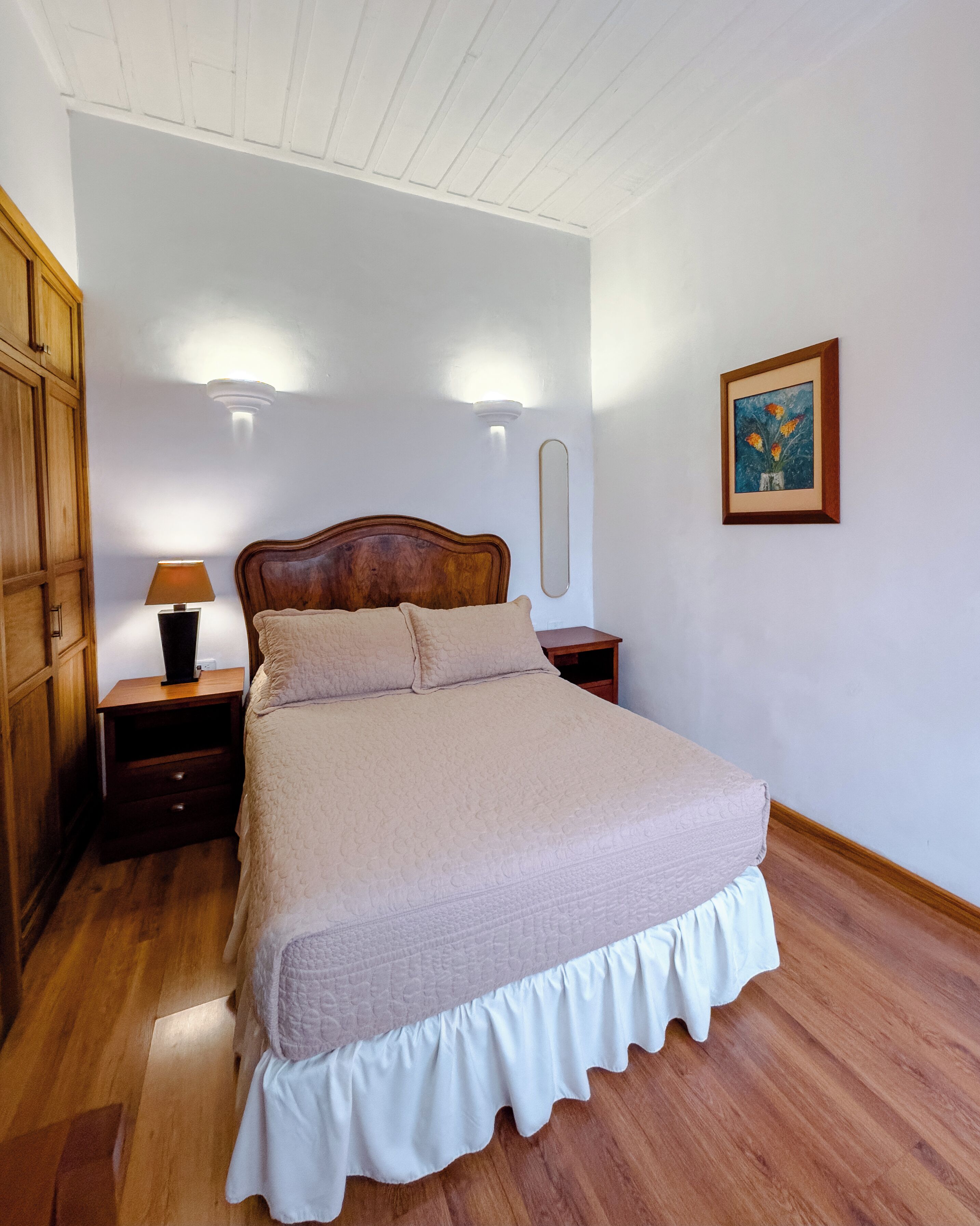 A bedroom with a wooden bed, a nightstand with a lamp, a dresser, and a framed picture on the wall.