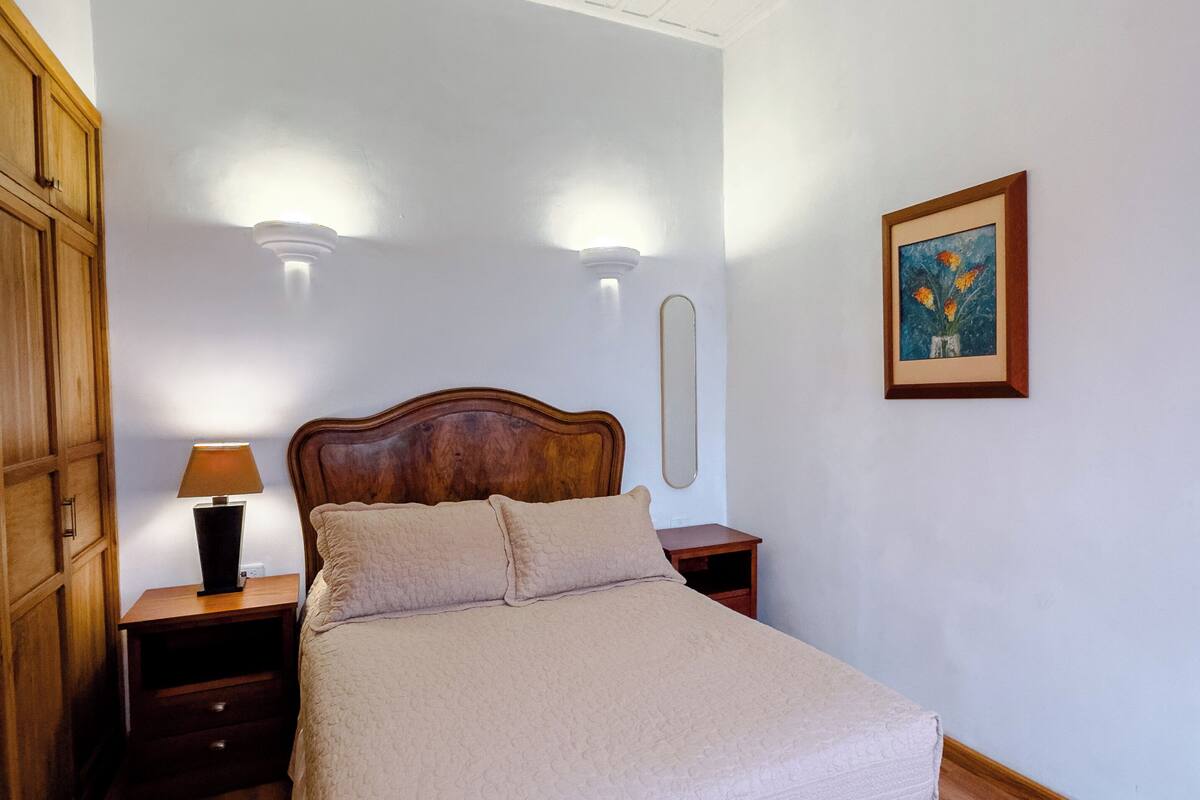 A bedroom with a wooden bed, a nightstand with a lamp, a dresser, and a framed picture on the wall.