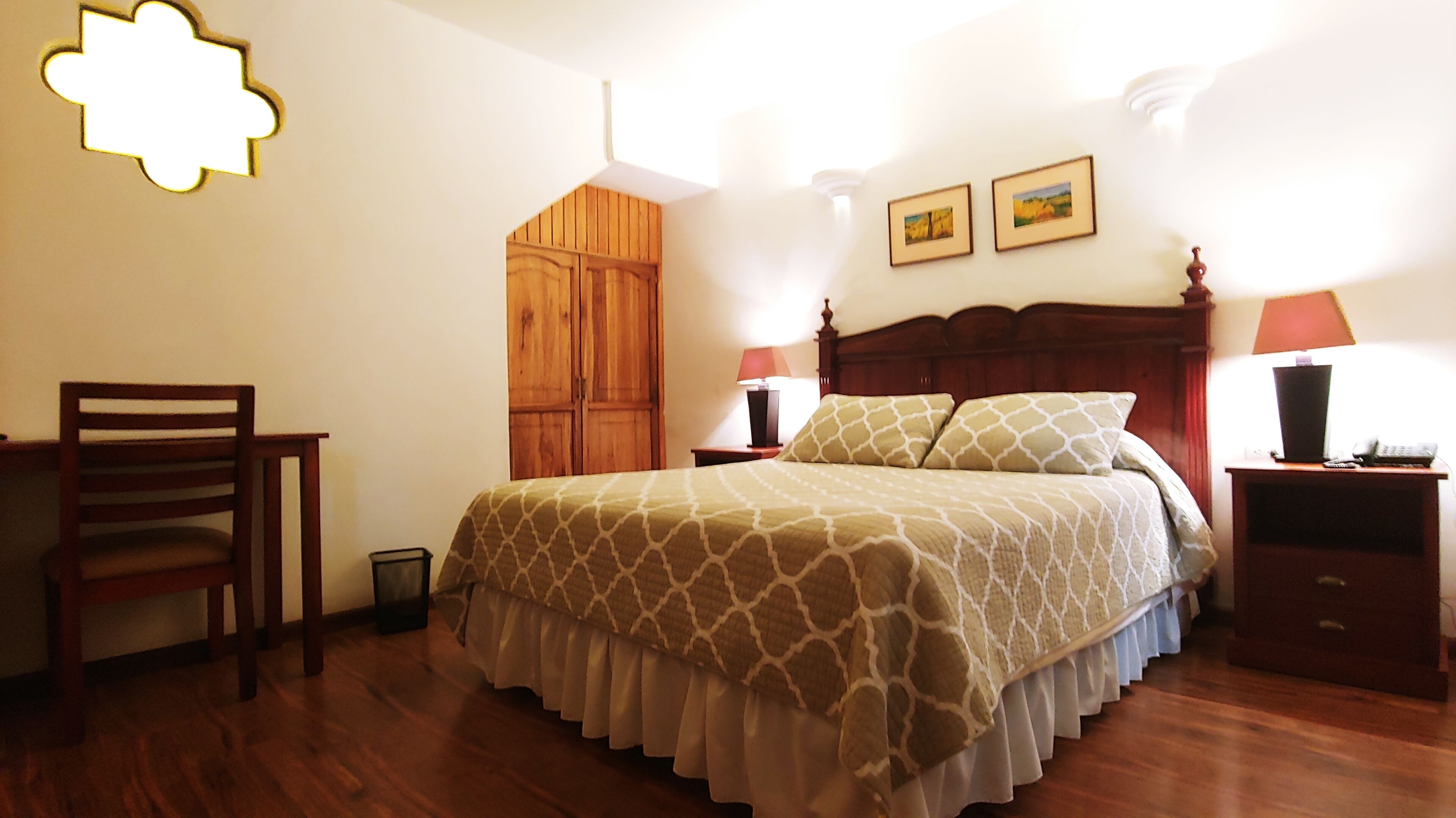 A bedroom with a wooden bed, a desk, a chair, a wooden wardrobe, and two framed pictures on the wall.