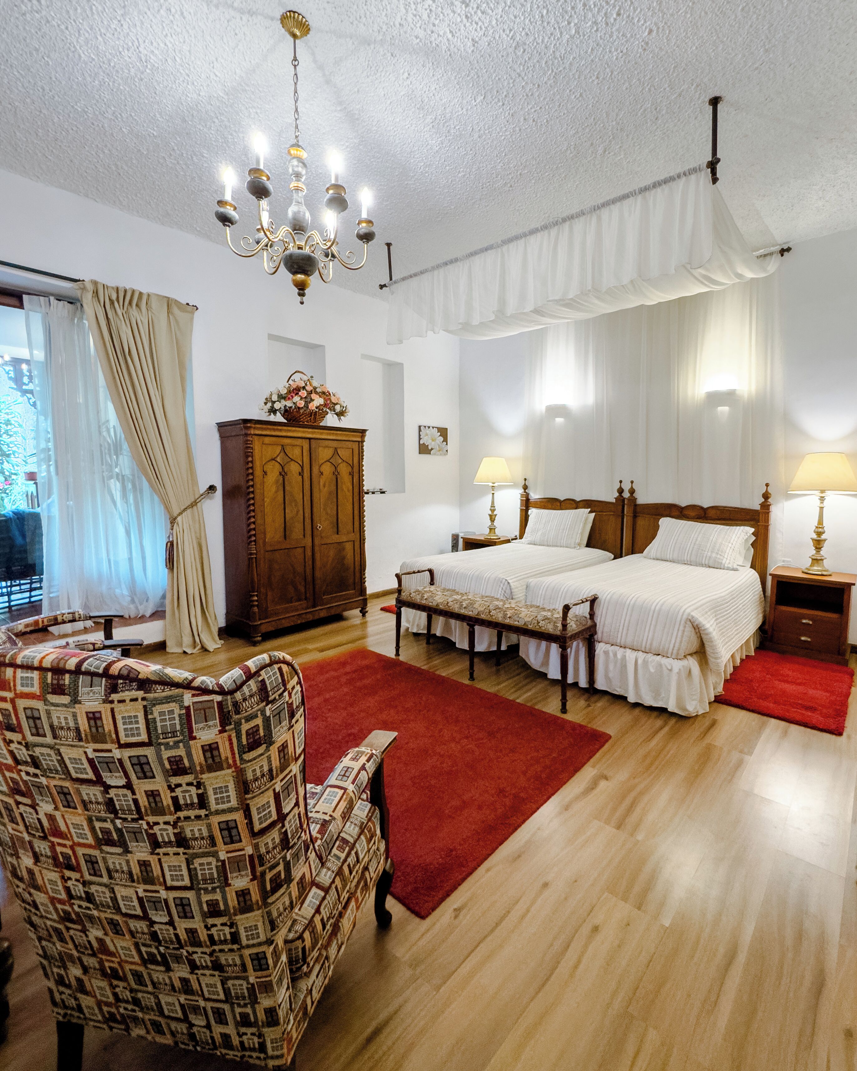 A bedroom with two beds, a wooden wardrobe, a patterned armchair, a red rug, and a chandelier.
