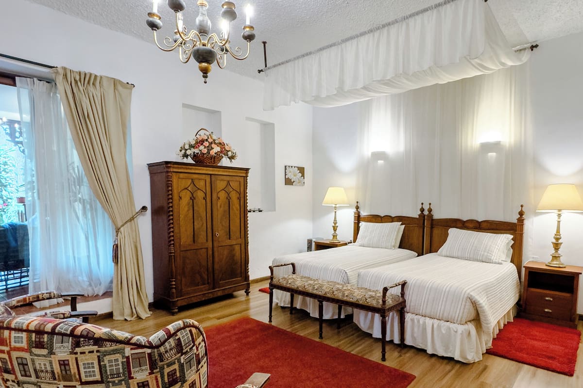 A bedroom with two beds, a wooden wardrobe, a patterned armchair, a red rug, and a chandelier.