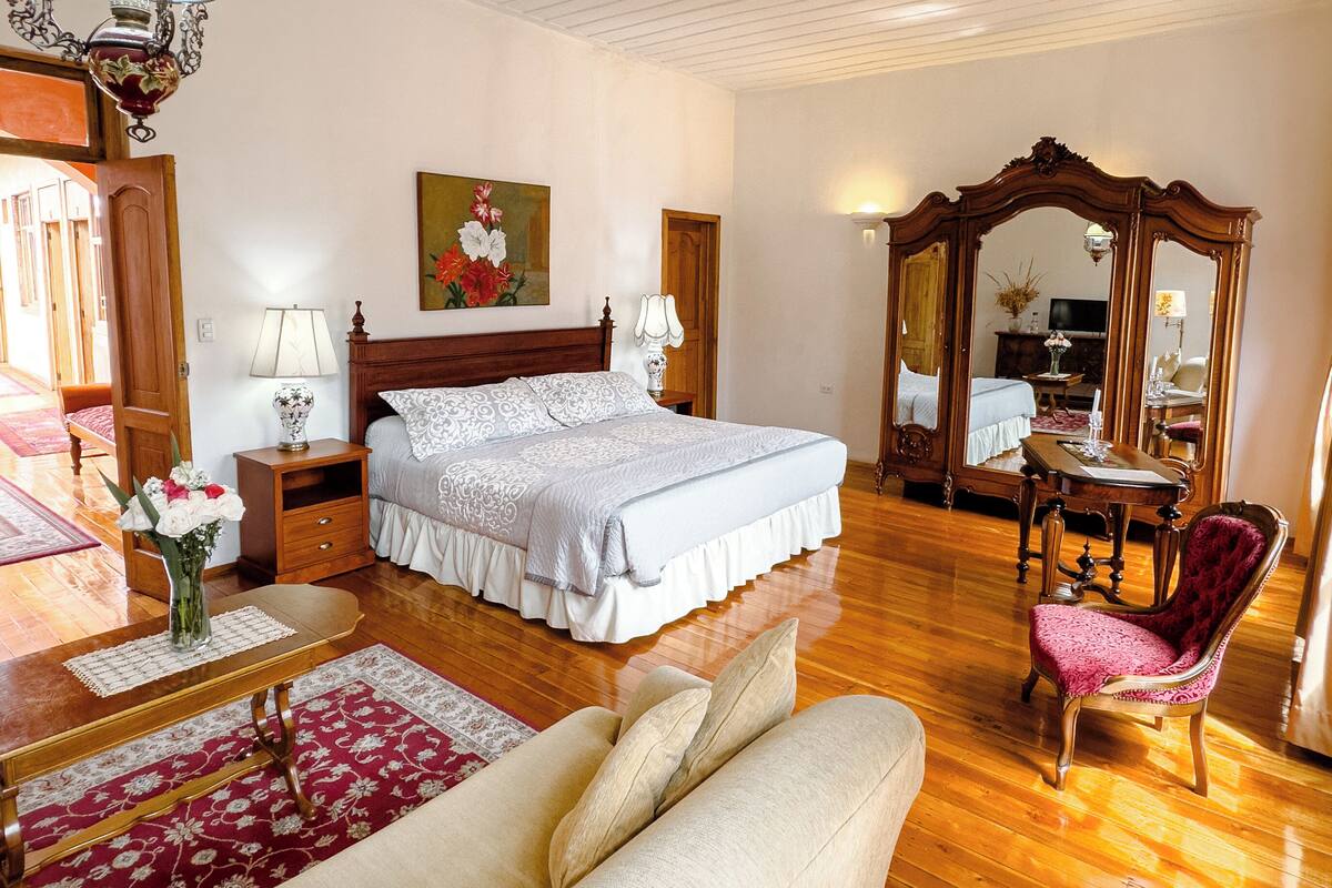 A spacious bedroom with a large bed, a sitting area with a sofa, a wooden floor, and a mirror.