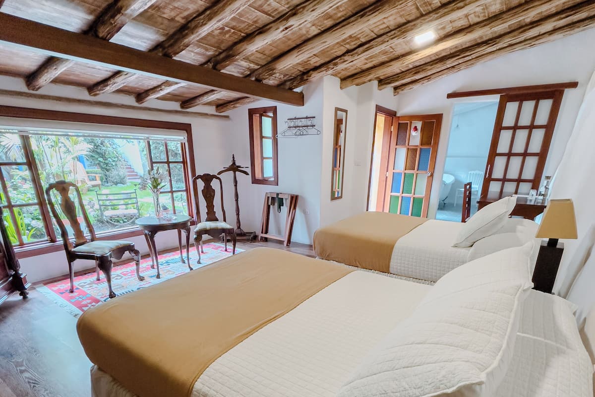 A bedroom with a large bed, wooden ceiling beams, a window with a view of greenery, and a small table with a chair.