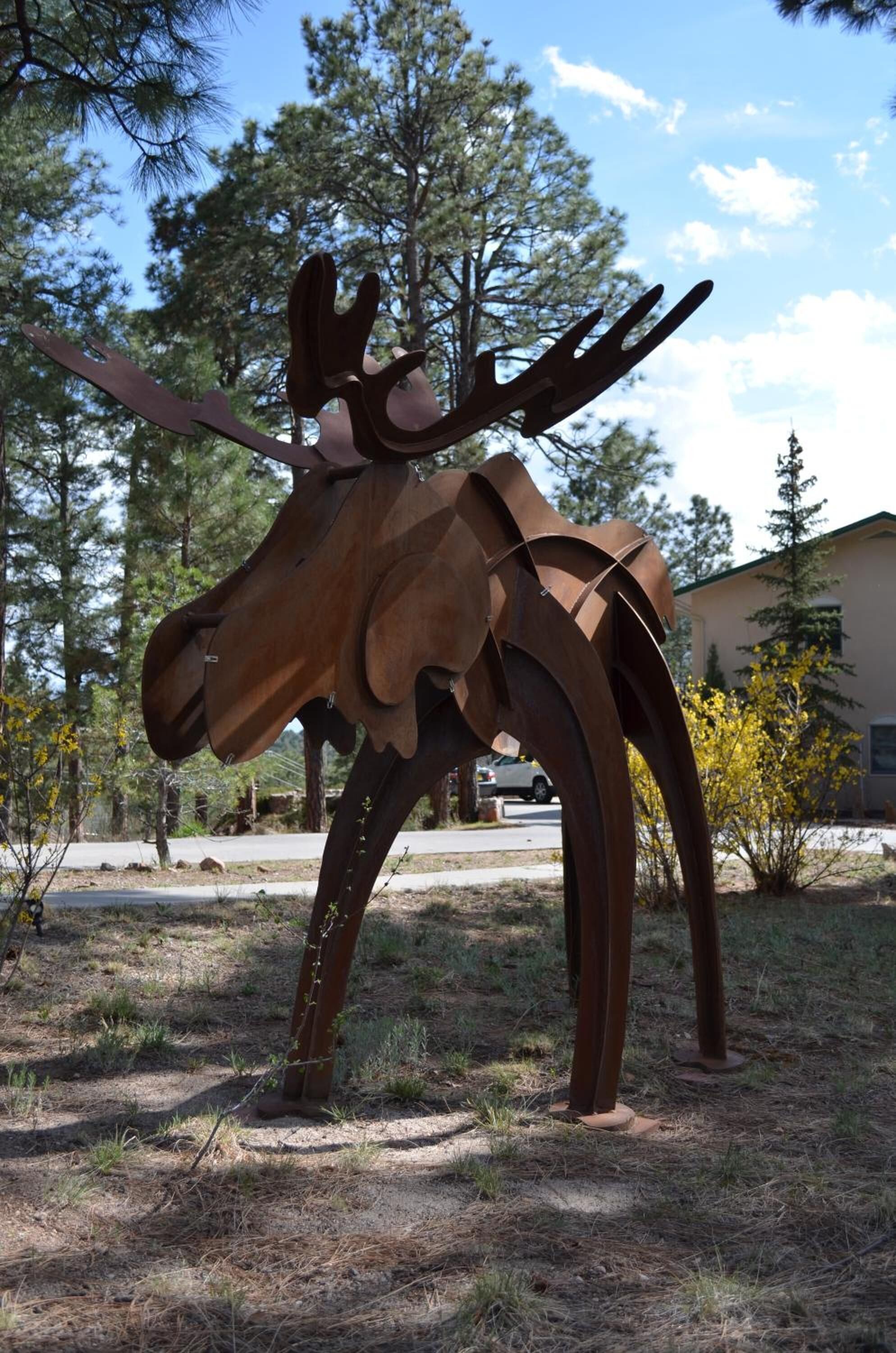 Una gran escultura de metal de un alce en un entorno exterior con árboles y un edificio al fondo.