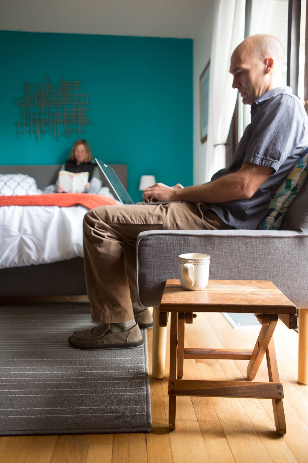 A modern room with a bed, a laptop on the bed, a coffee mug on a wooden table, and a person sitting on the bed.