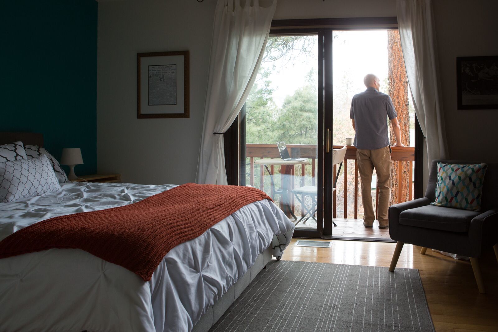 A bedroom with a bed, a red blanket, a chair, and a balcony with a person standing.