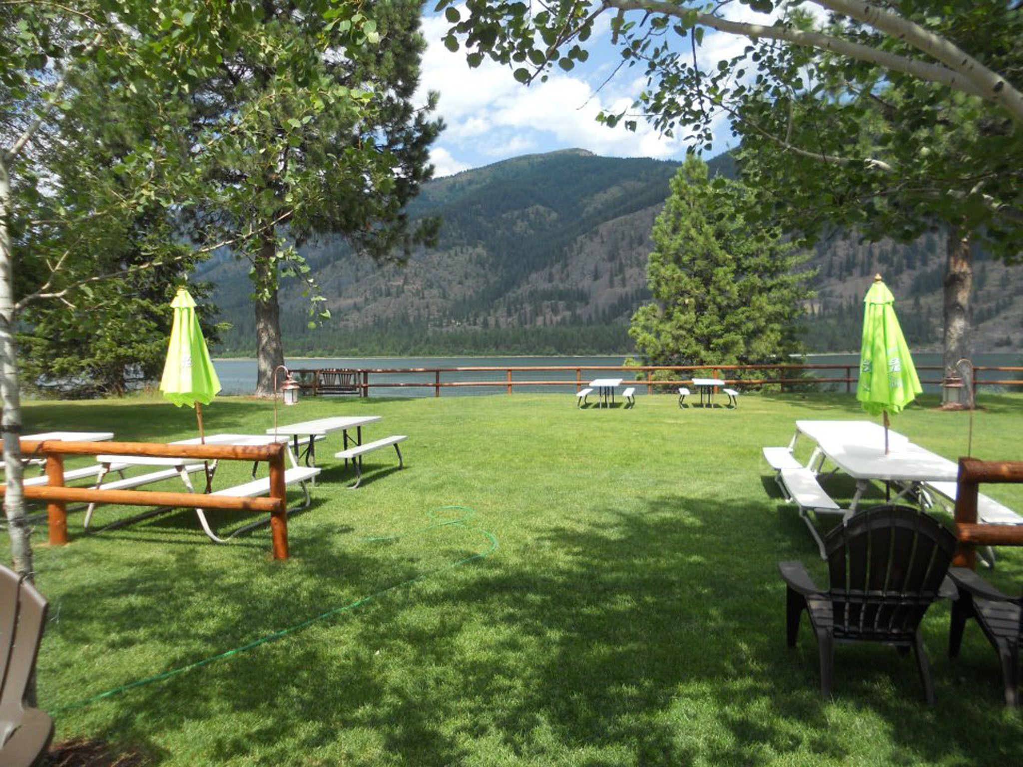 Un espace pique-nique équipé de tables et de chaises, entouré d’une clôture en bois, et offrant une vue sur un lac et des montagnes.