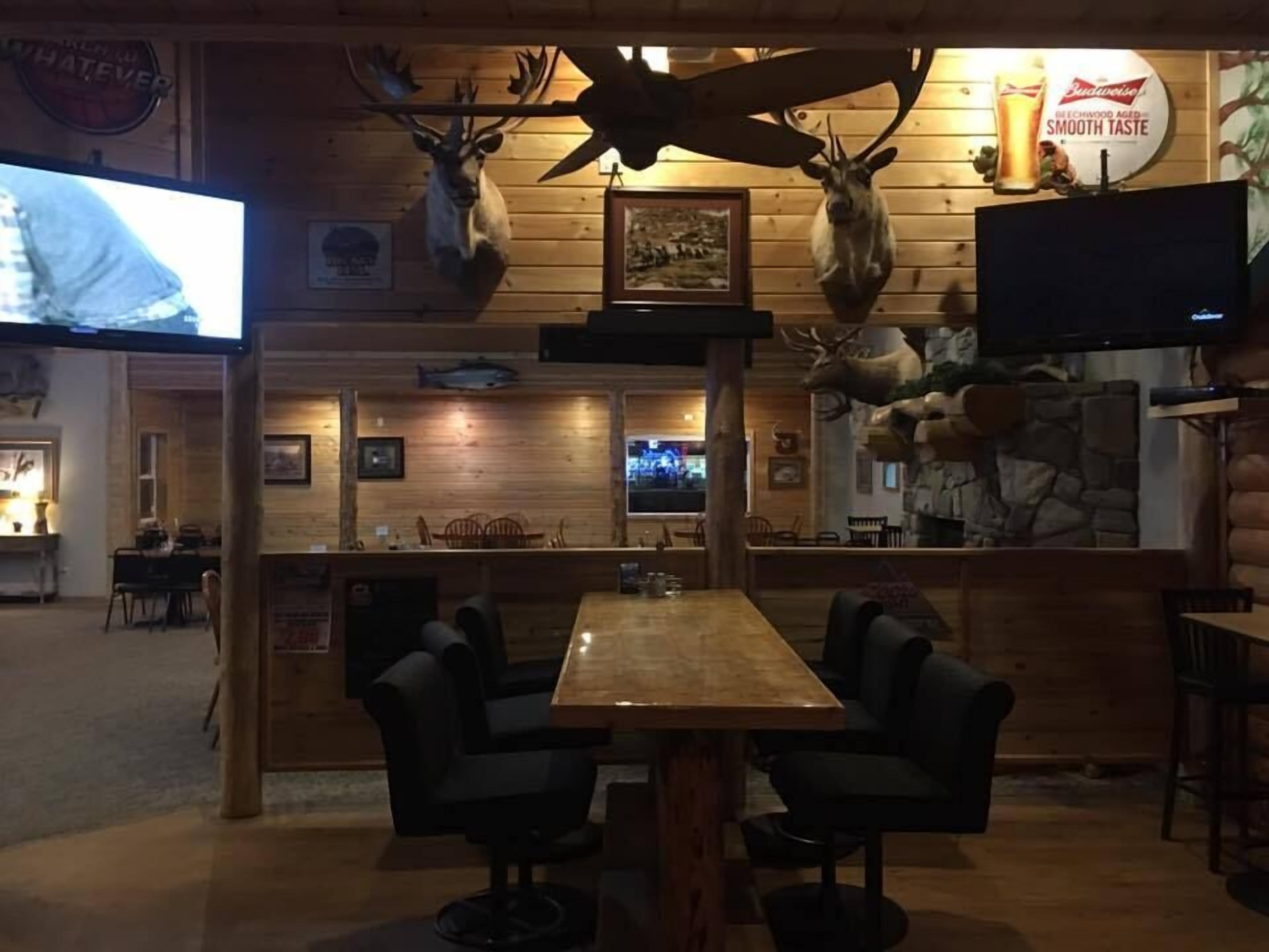 A rustic interior with wooden walls, mounted animal heads, a television, and a bar area with seating.
