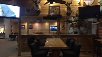A rustic interior with wooden walls, mounted animal heads, a television, and a bar area with seating.