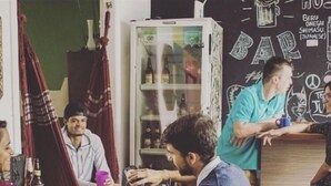 Un groupe de personnes est assis autour d’une table dans un espace de restauration informel. On aperçoit un réfrigérateur rempli de boissons et un mur décoré de différents objets.