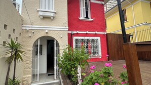 Une maison à deux étages avec une façade rouge, des volets blancs et un petit jardin agrémenté de fleurs roses.