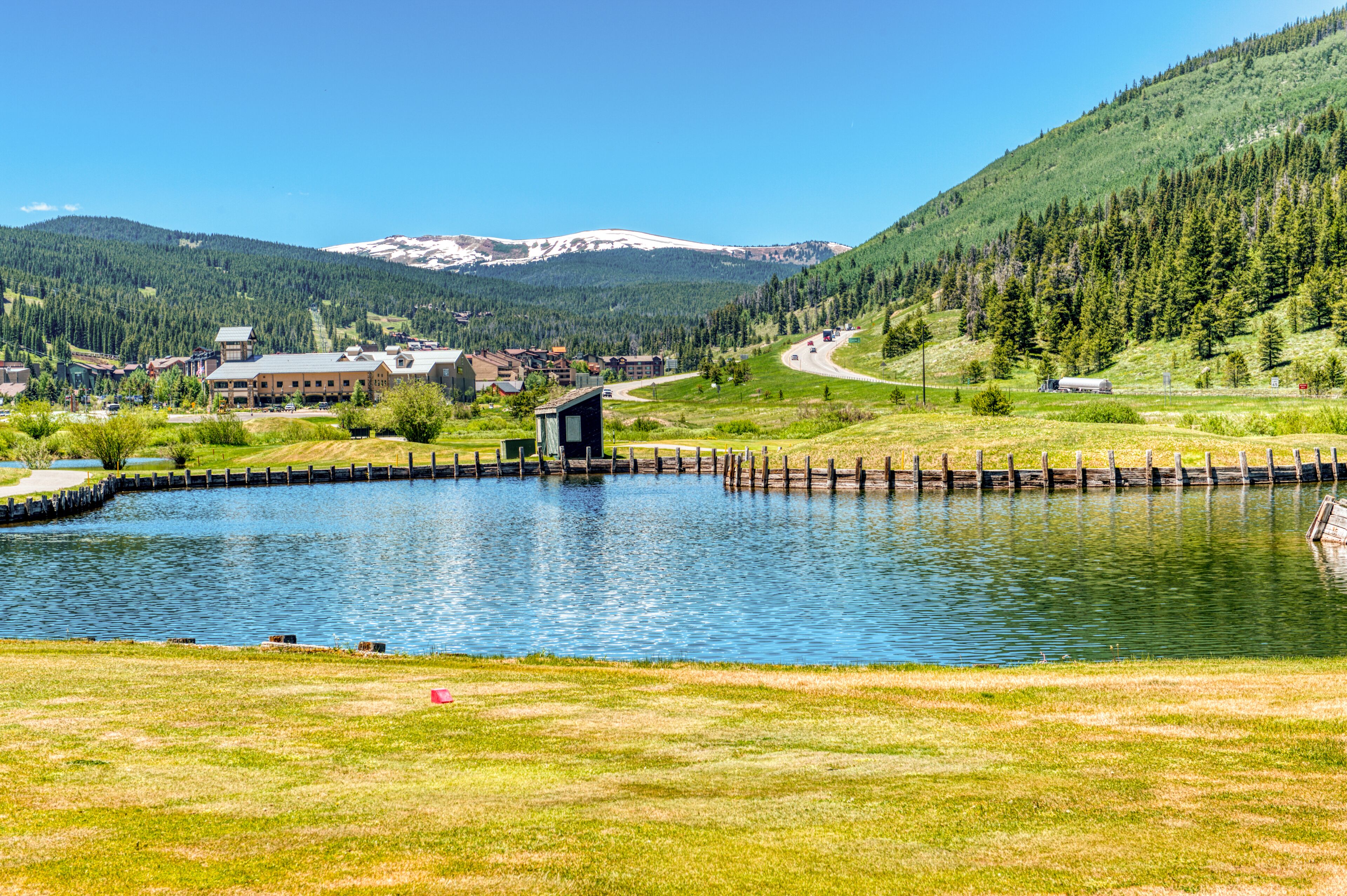 Vista para o lago com um pequeno cais, edifícios ao fundo e uma paisagem montanhosa.