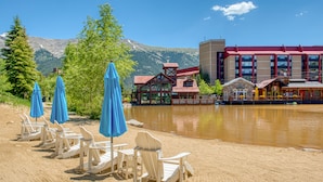 Um resort à beira do lago com cadeiras de madeira e guarda-sóis azuis, um edifício com telhado vermelho e montanhas ao fundo.