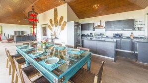 A spacious dining area with a long table set for four, a kitchen with modern appliances, and a high ceiling with wooden beams. - Baan Ling Noi Villa (Koh Samui)