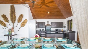 A dining area with a glass table set for six, surrounded by wicker chairs. A ceiling fan is visible above, and a kitchen with dark cabinets and a stove is in the background. - Baan Ling Noi Villa (Koh Samui)