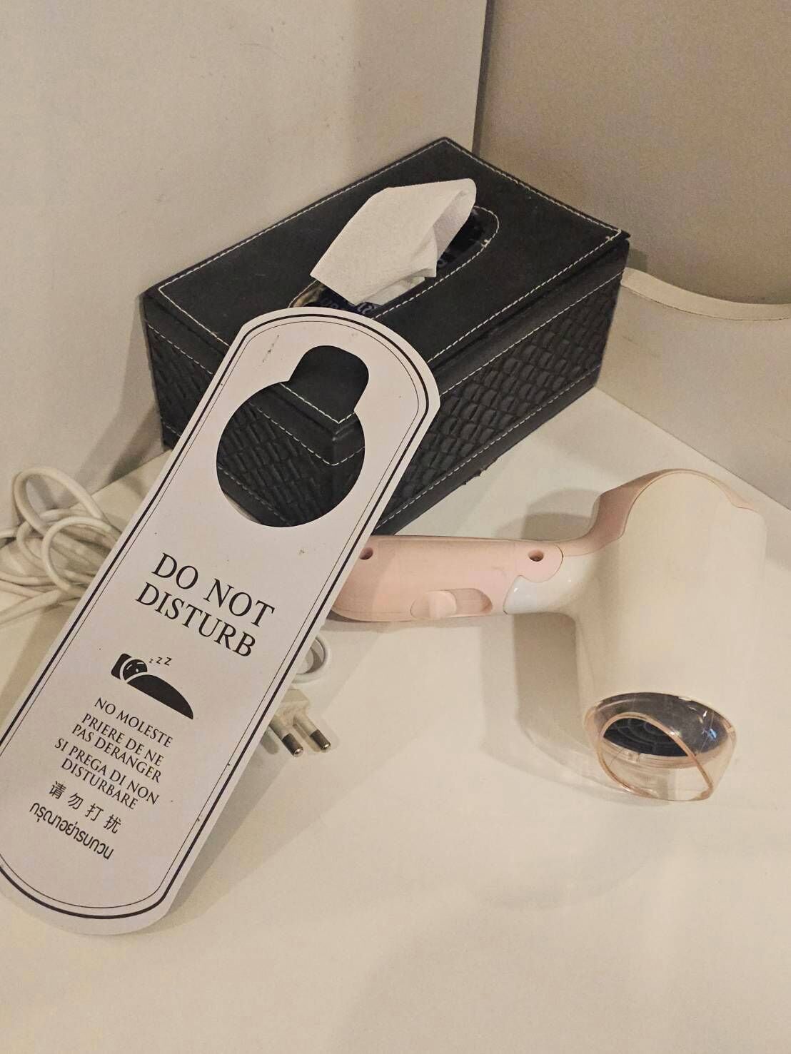 A hair dryer with a 'DO NOT DISTURB' sign, a tissue box, and a cup on a white surface.