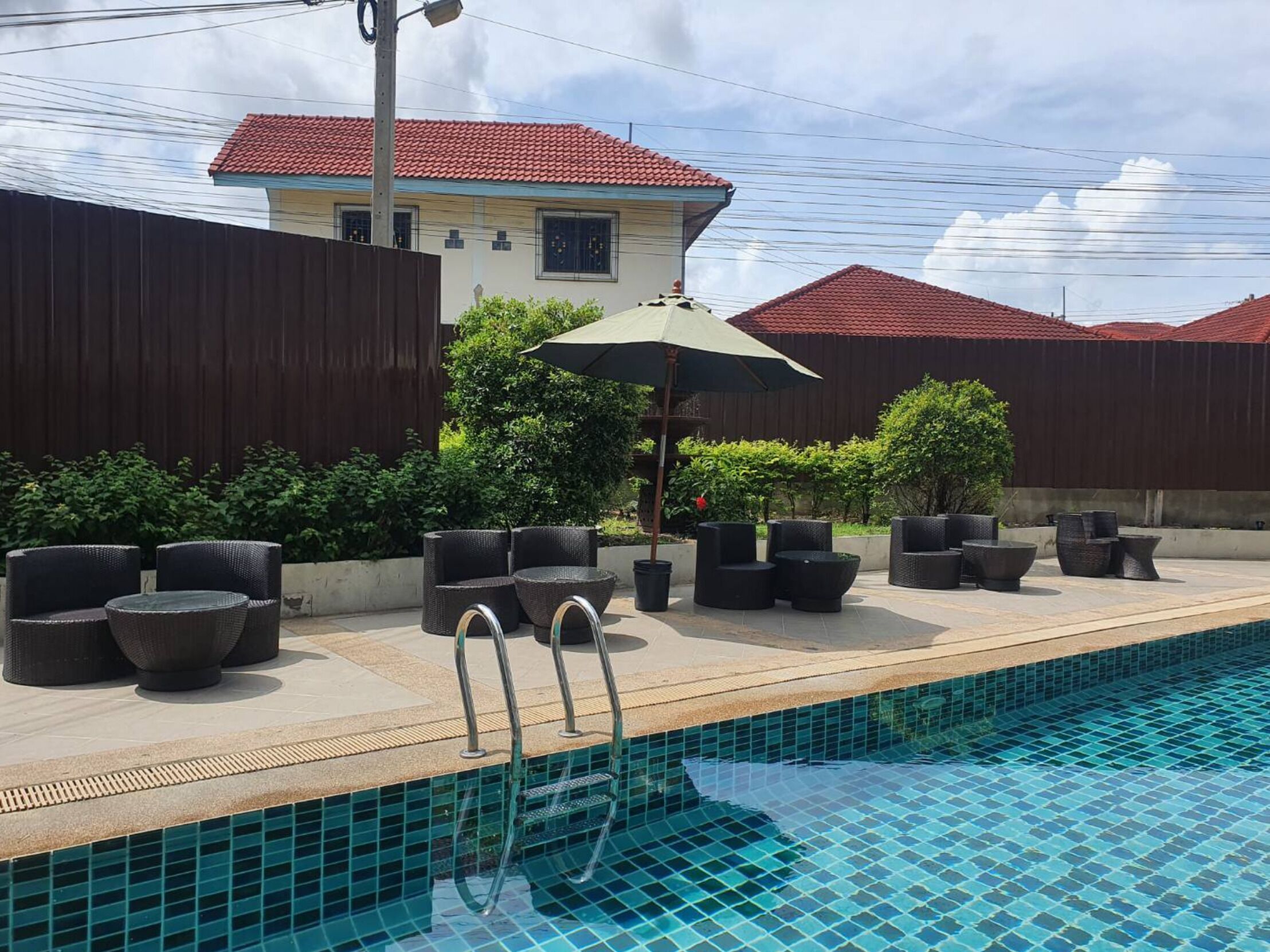 A swimming pool with lounge chairs and an umbrella, surrounded by a wooden fence and a building with a red roof.