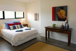 A neatly made bed with a white comforter and blue and red pillows, a bedside table with a lamp, and a painting on the wall.