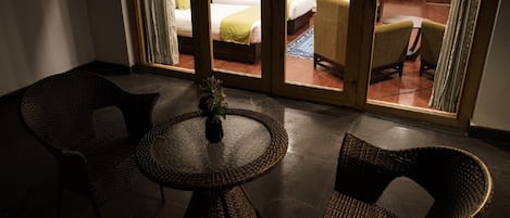 A room with a round table and two wicker chairs, a small vase with flowers, and a view of a bedroom with a bed and a chair through a glass door.