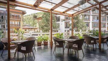 A covered outdoor seating area with wicker furniture and potted plants, overlooking a mountainous landscape.