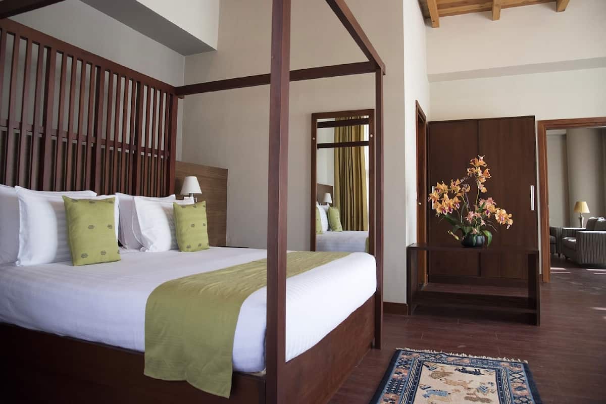 A four-poster bed with a wooden headboard, a green throw blanket, and multiple pillows. A vase with flowers is on a side table next to the bed.