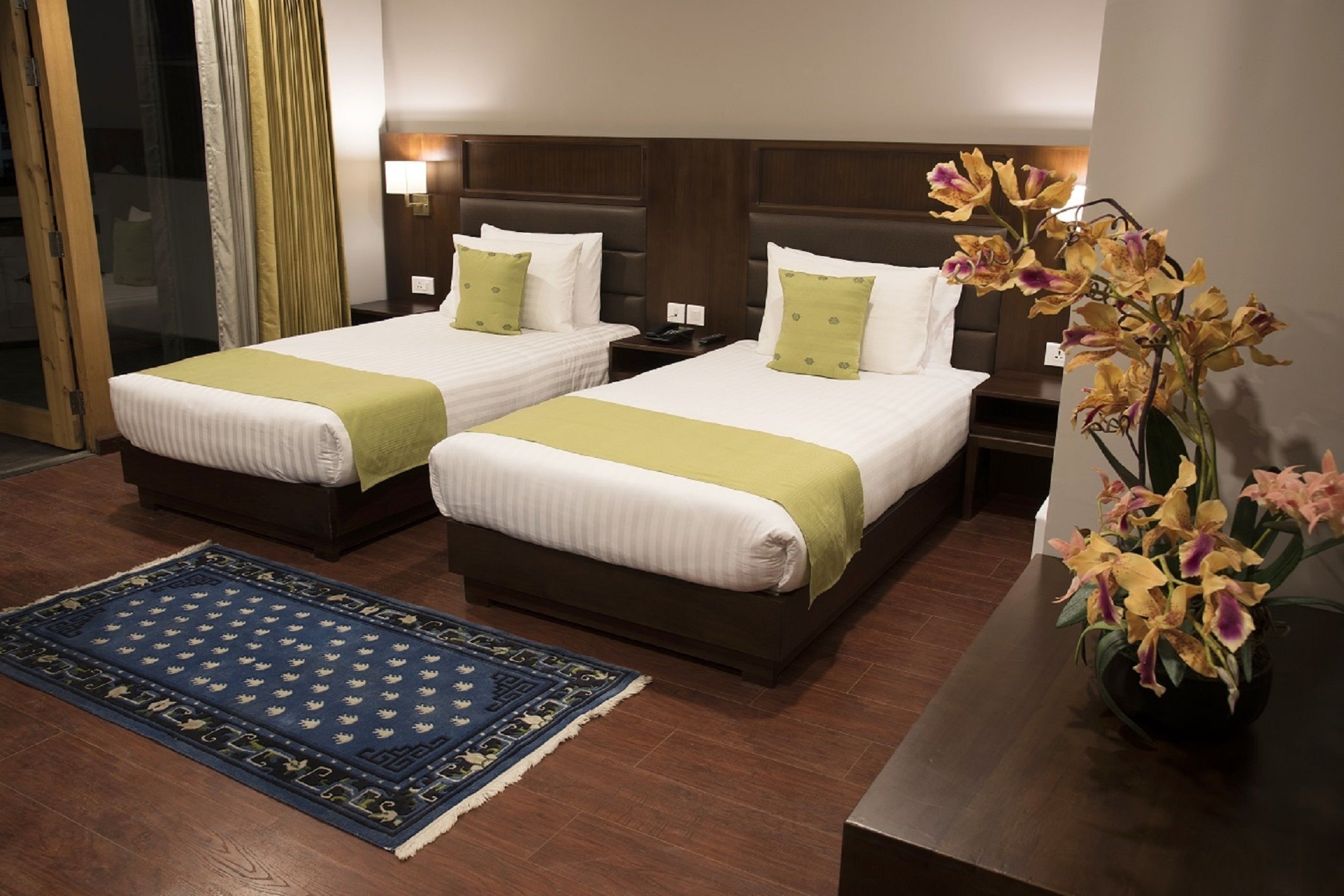 Habitación de hotel con dos camas, un tapete azul estampado y un jarrón con flores.