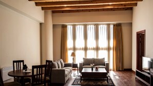 Sala de estar con sofá, mesa de centro y sillas. Un televisor está a la derecha y una ventana con cortinas.