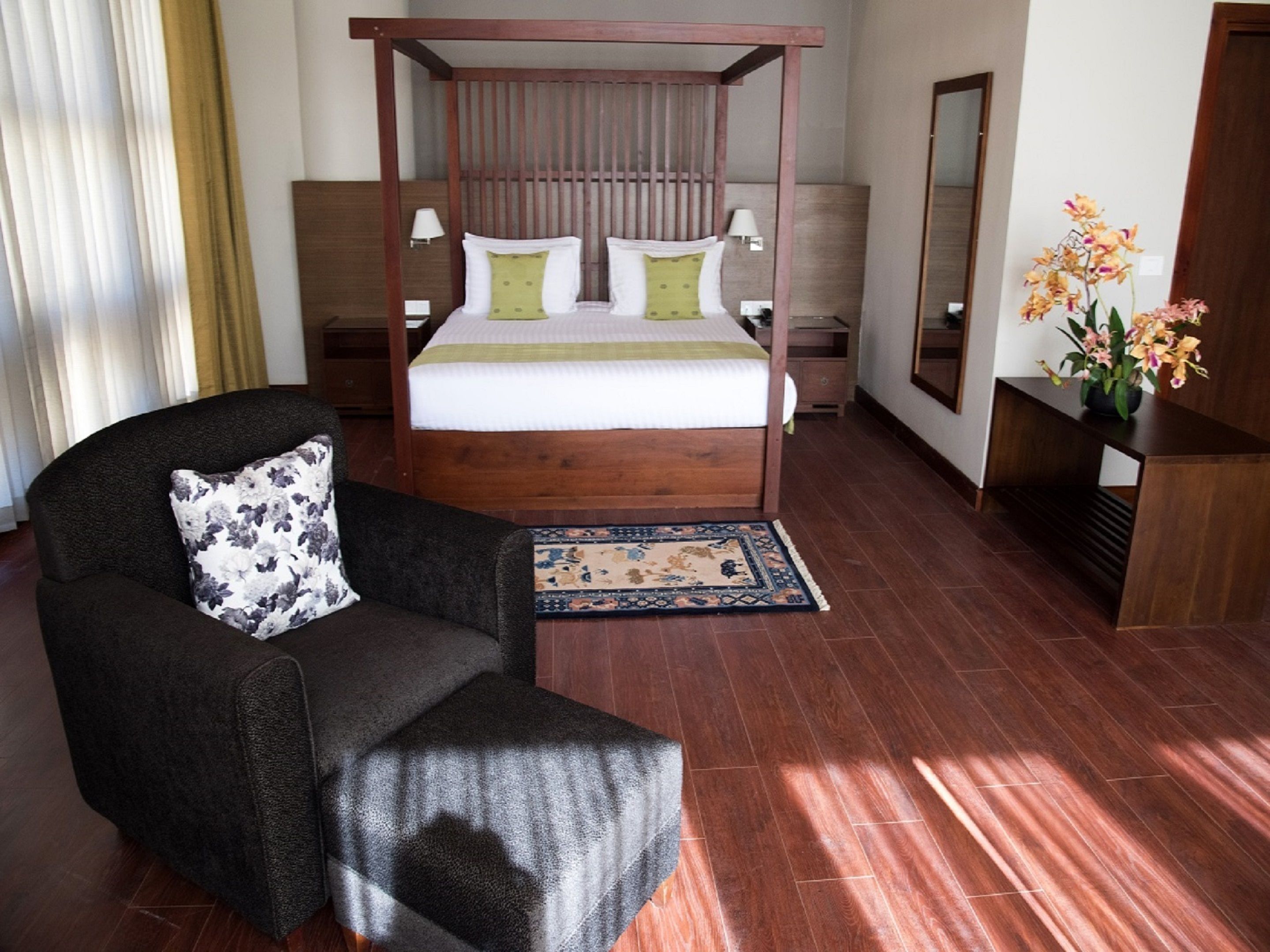 Una cama con dosel de madera, dos mesitas de noche, un sillón oscuro con un cojín floral, una alfombra pequeña y una mesita de madera con un jarrón de flores.