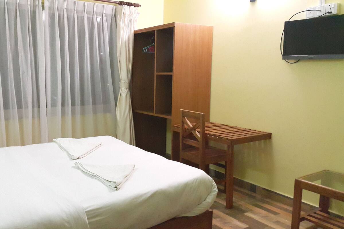 Una cama bien hecha con una colcha blanca y dos toallas dobladas. Una silla de madera y una mesita están ubicadas junto a la cama. Un armario de madera se encuentra apoyado contra la pared.
