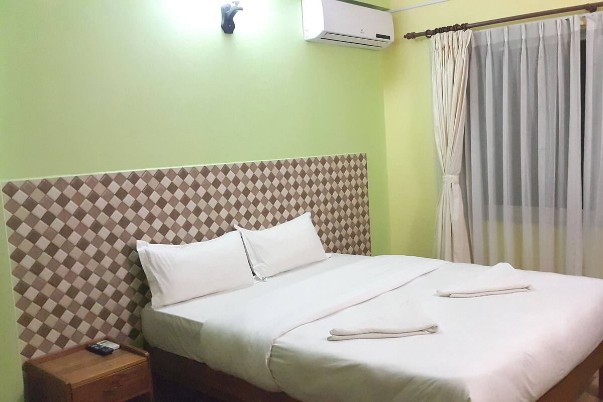 Una cama bien hecha con sábanas y almohadas blancas, una mesita de noche de madera con un control remoto, un cabecero estampado y una ventana con cortinas traslúcidas.