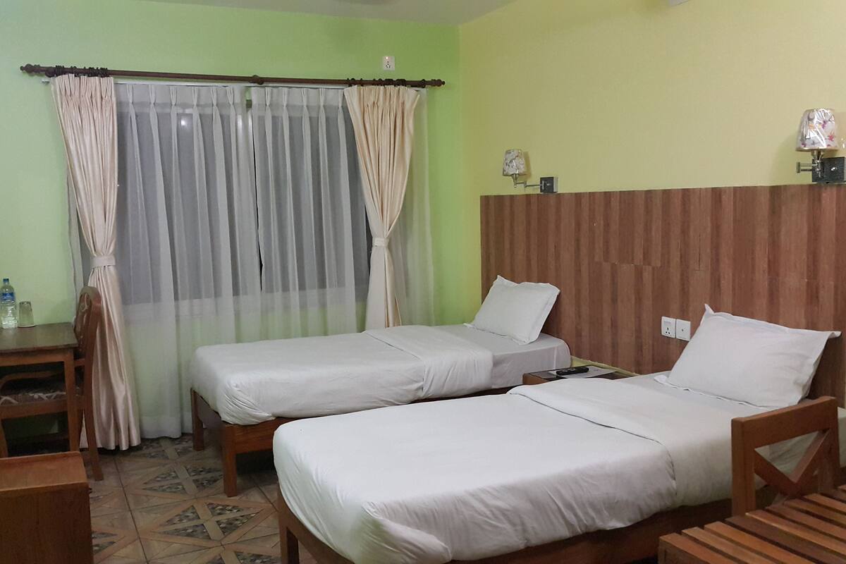 Habitación de hotel con dos camas individuales, un pequeño escritorio de madera, una silla, una mesita de noche, una botella de agua y una lámpara.