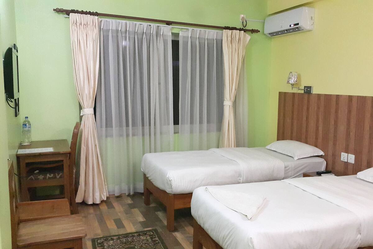 Una habitación de hotel con dos camas individuales, un cabecero de madera, una mesita con una silla, una botella de agua y un aire acondicionado instalado en la pared.