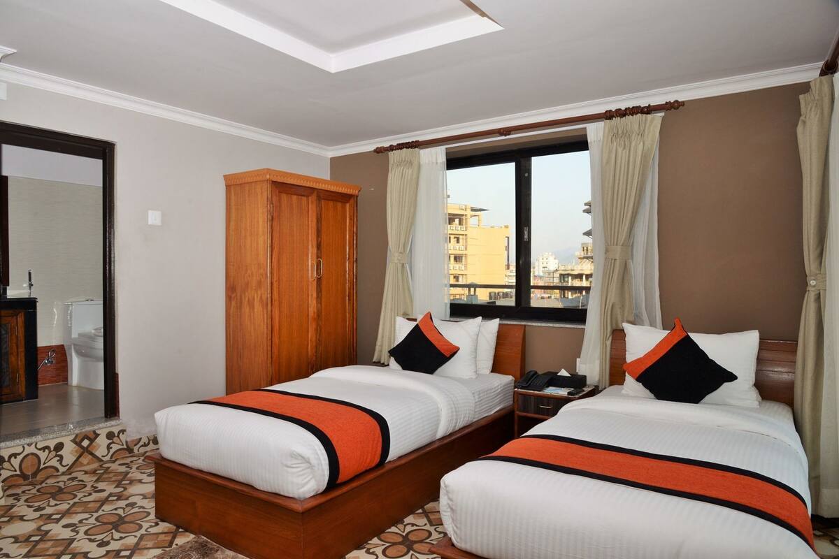 A hotel room with two beds, a wooden wardrobe, a window with curtains, and a view of buildings outside.