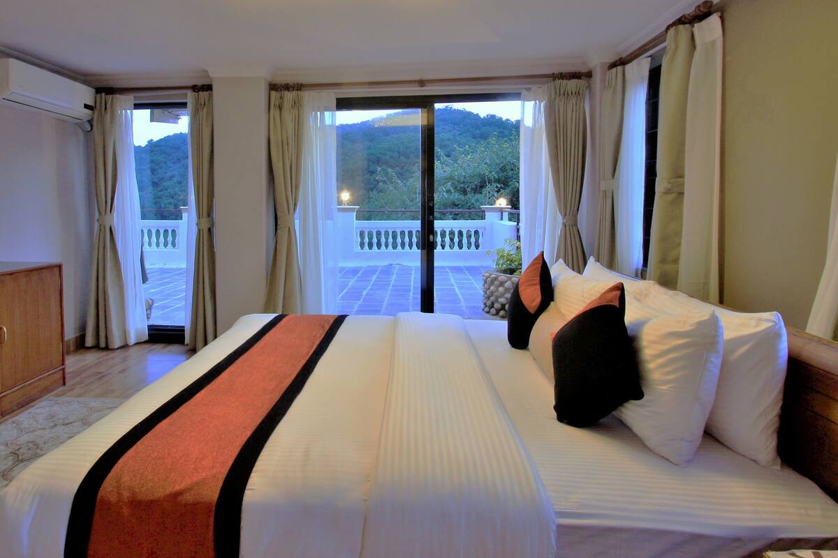 A hotel room with a bed, curtains, and a view of a balcony with a railing and greenery.