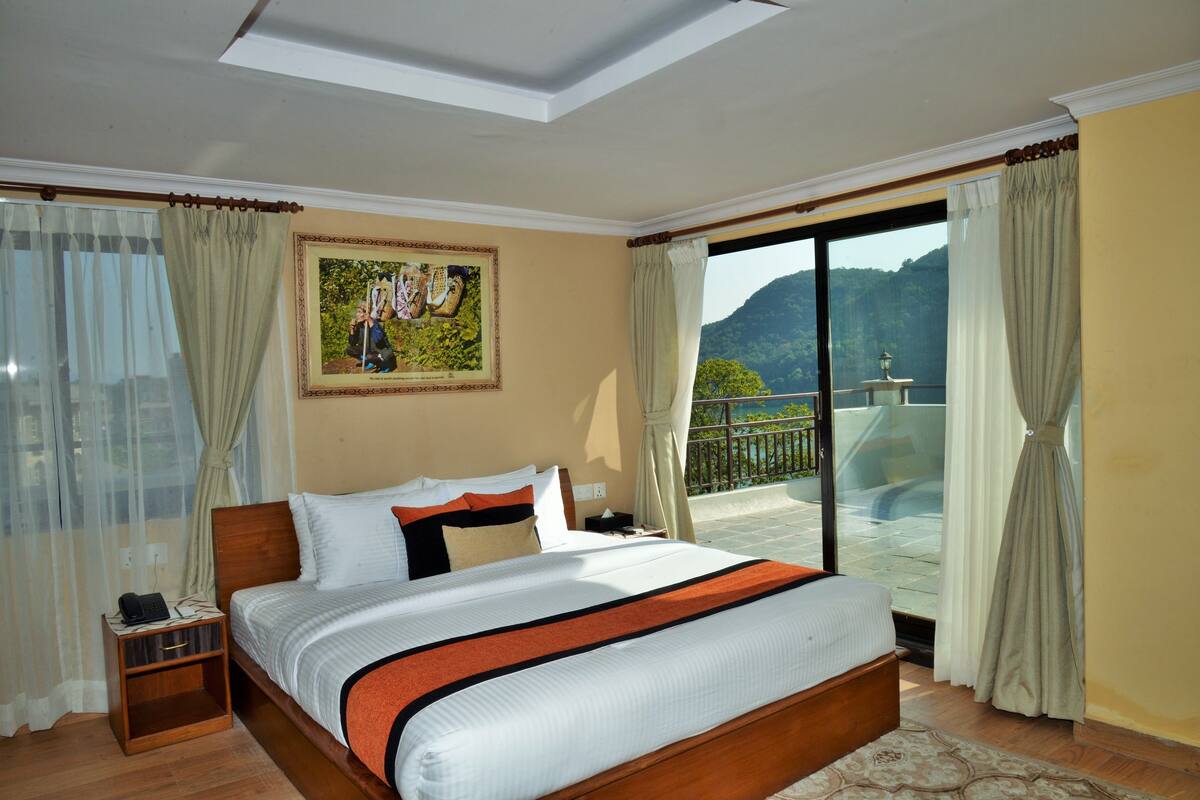 A hotel room with a large bed, a nightstand, a painting on the wall, and a balcony with a view of the mountains.