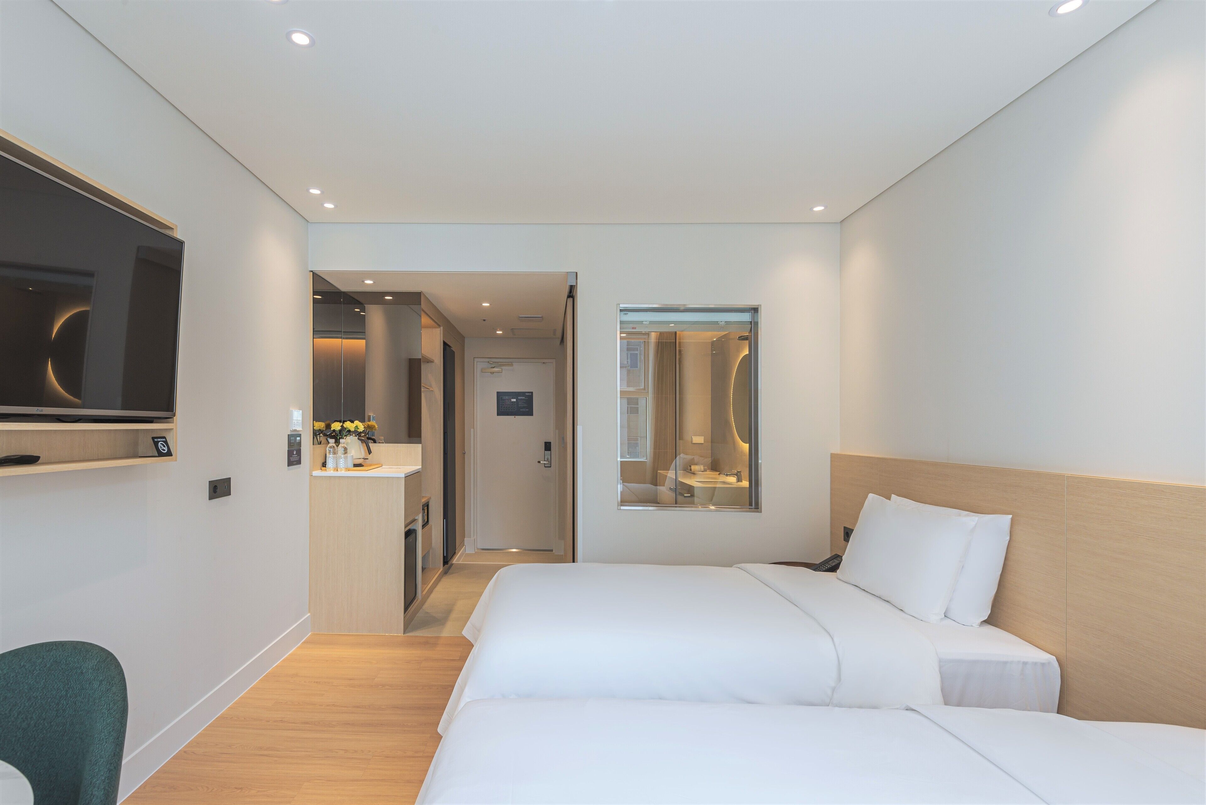 Habitación de hotel con dos camas, un televisor montado en la pared, una kitchenette pequeña y un baño visible a través de una puerta de vidrio.