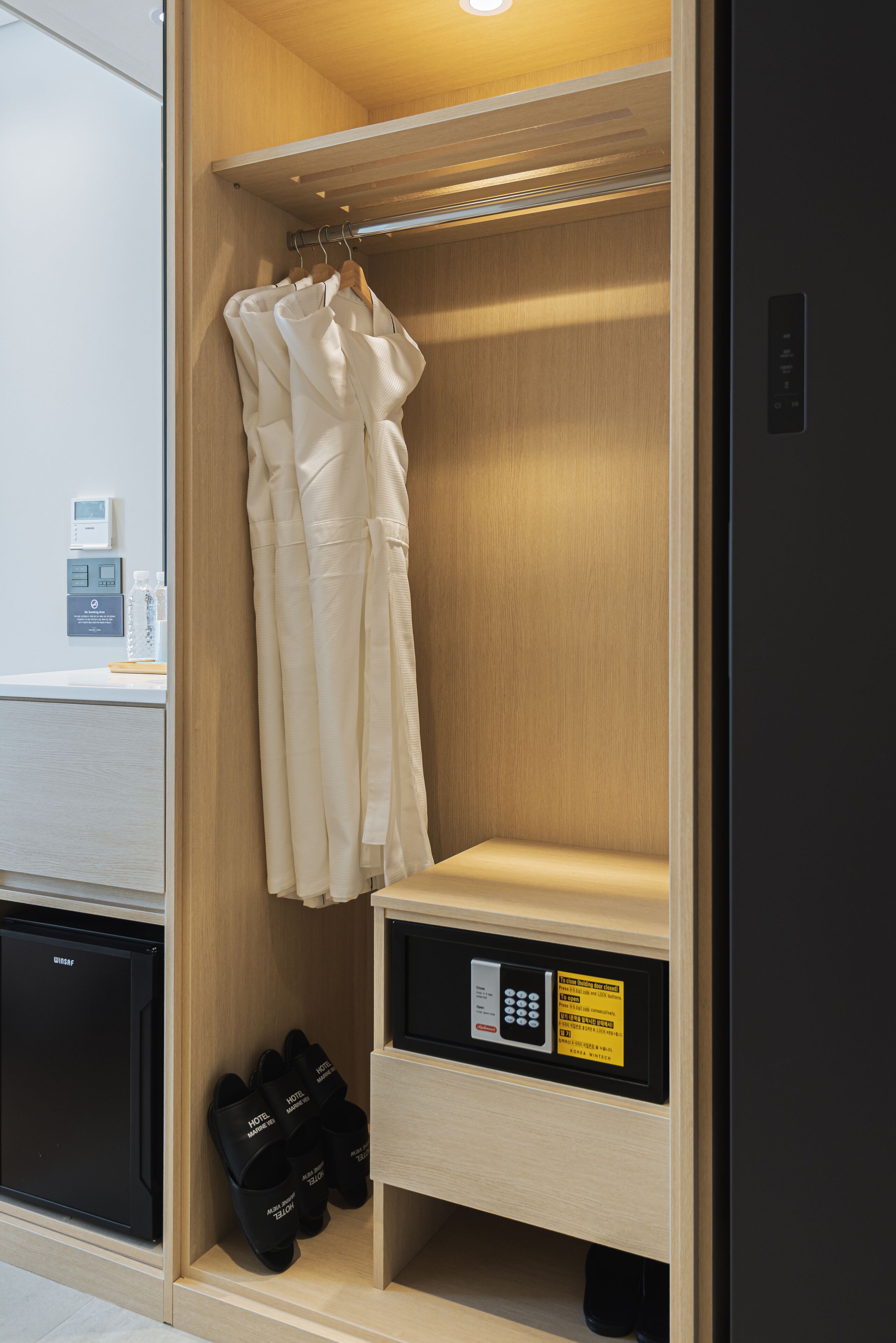 A hotel room with a wooden wardrobe, a hanging robe, and a shoe rack with black slippers.