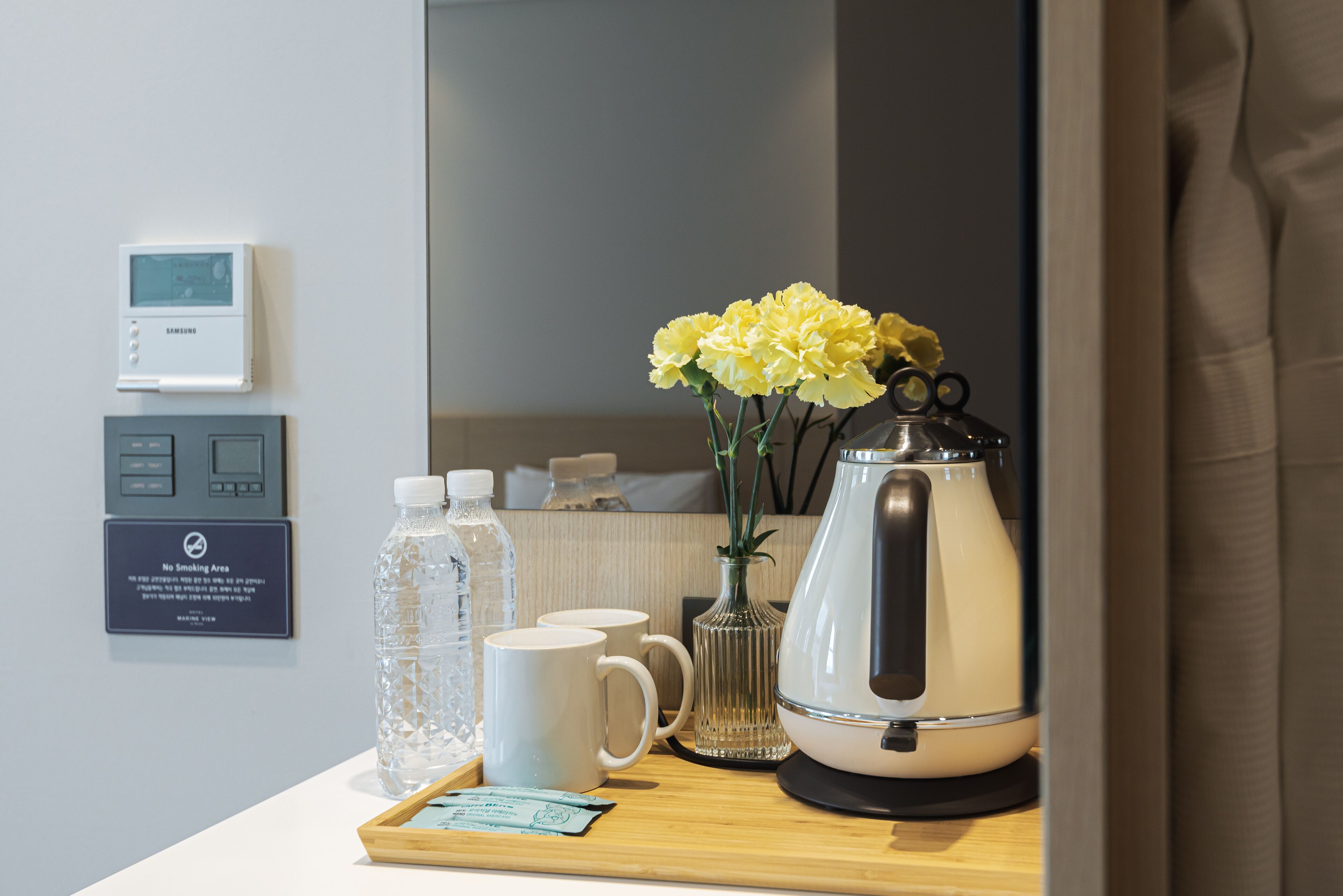 Habitación de hotel con termostato digital, un cartel de 'No Fumar', una botella de agua, dos tazas, un florero con flores y una cafetera sobre una bandeja.