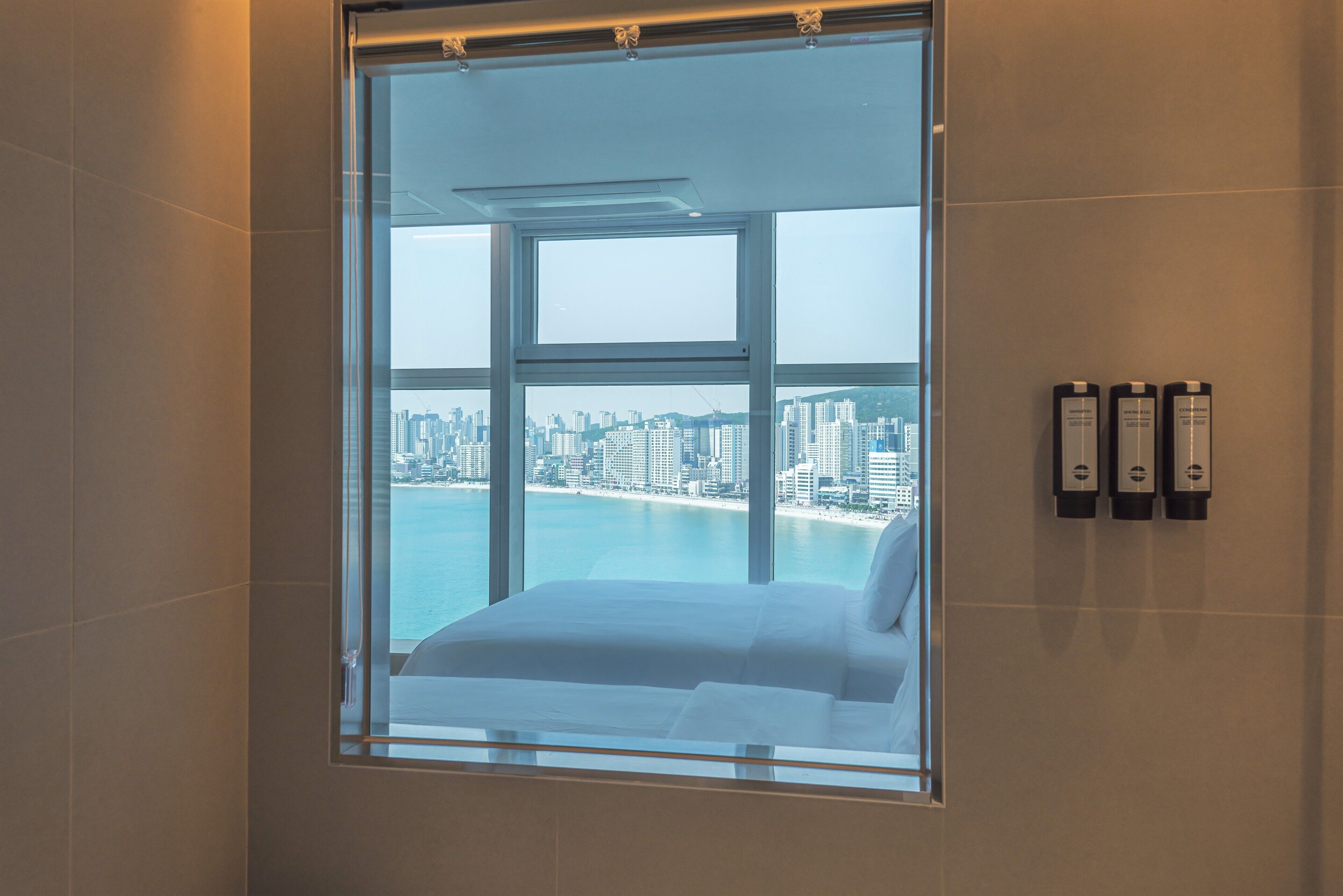 A hotel room with a large window offering a cityscape view, a bed, and three wall-mounted air conditioning units.
