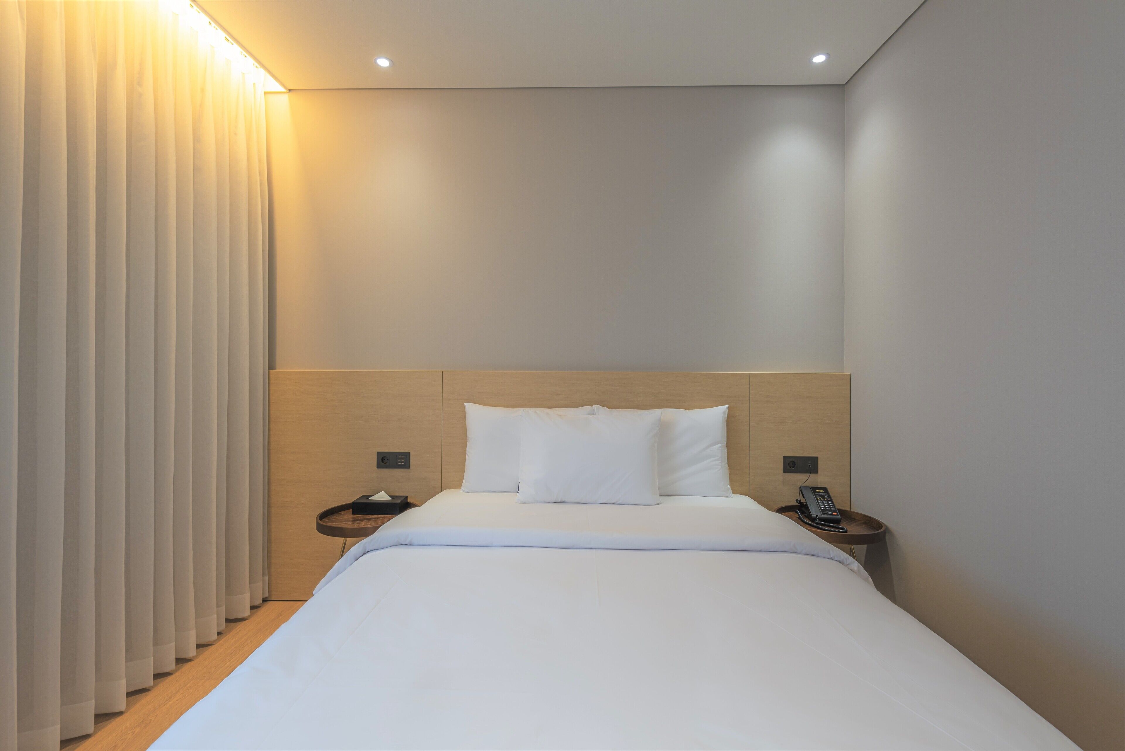 Una habitación de hotel moderna con una cama bien hecha, una mesita de noche con un teléfono y una lámpara fijada en la pared.