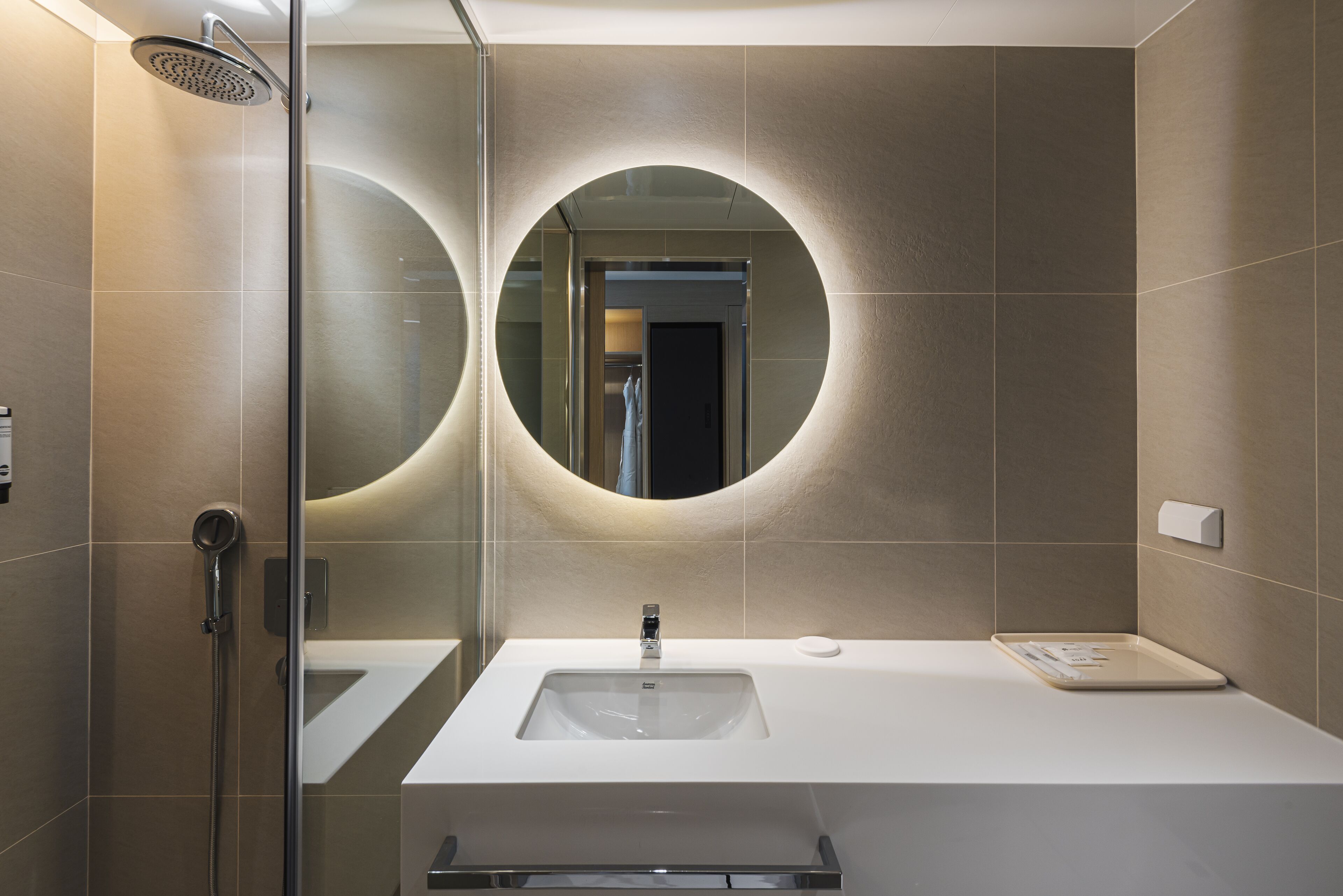 Une salle de bain moderne avec une douche à parois de verre, un grand miroir à cadre rond et un lavabo blanc doté d’un porte-savon.