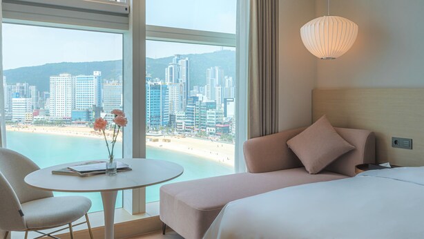 Een hotelkamer met een bed, een bank, een tafel met een bloevas en uitzicht op een strand en stadsgezicht.