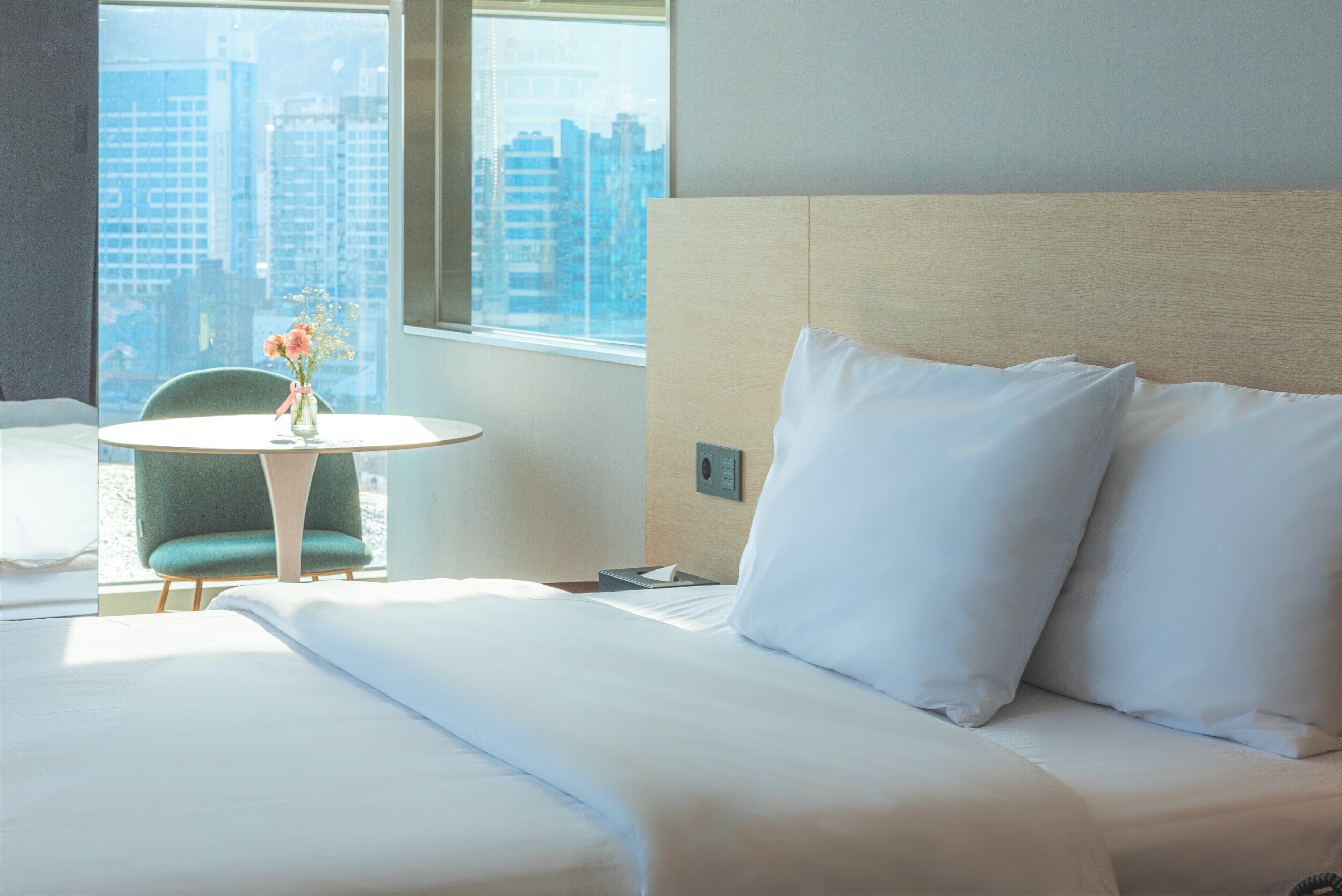 Ein Hotelzimmer mit einem Bett, einem Sessel, einem kleinen Tisch mit Blumenvase und Blick auf Stadtgebäude durch das Fenster.